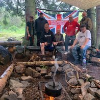 A group of people camping in a forest in front of a log fire with a kettle